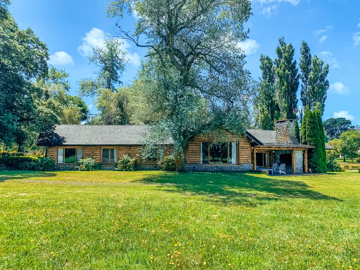 La casa de troncos entre álamos y árboles añosos, con el parque de césped al frente